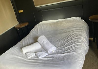 A cozy bedroom in Cabin 54 at Stawell Hotel Motel Accommodation featuring a neatly made double bed with fresh white linens and neatly rolled towels. The room is illuminated by natural light streaming through the window, which is adorned with blinds for privacy. Two round bedside tables flank the bed, providing a stylish and comfortable space for guests to relax