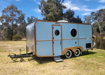 A charming blue caravan situated by a serene body of water, surrounded by lush greenery at Stawell Hotel Motel Accommodation. The caravan features a sleek design with wooden accents, including a circular window and a welcoming entrance. Its compact size is perfect for camping enthusiasts seeking a cozy getaway. The grassy area around the caravan provides an ideal space for relaxation and outdoor activities, enhancing the overall inviting atmosphere of this picturesque setting
