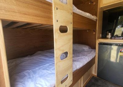 Inside view of the blue caravan at Stawell Hotel Motel Accommodation, showcasing a cozy sleeping area with two neatly made bunk beds. The wooden structure features a light wood panel serving as a partition between the bunks, enhancing safety and privacy. A compact refrigerator can be seen in the corner, providing convenience for guests. The warm tones of the wood and the clean white bedding create a welcoming and comfortable atmosphere, perfect for relaxation after a day of exploring.