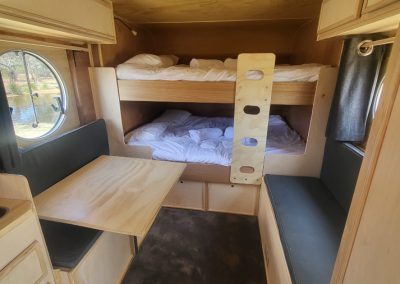 Interior of the blue caravan at Stawell Hotel Motel Accommodation, featuring a cozy sleeping space with a pair of bunk beds topped with white bedding. A practical wooden table is positioned beside the bunk beds, offering a space for dining or relaxation. The design includes cushioned seating along the walls, enhancing comfort. A large circular window lets in natural light, creating an inviting atmosphere. The wood paneling and warm colors contribute to a rustic yet modern charm, making it an ideal retreat for guests