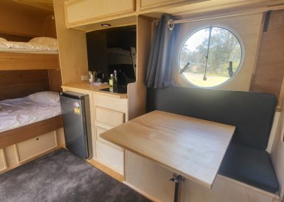 The interior of the blue caravan at Stawell Hotel Motel Accommodation showcases a well-designed living space. The image features a cozy nook with a small table that can be folded down for dining or working, surrounded by plush seating. Above, a set of bunk beds is neatly made with white linens, providing comfortable sleeping arrangements. A compact refrigerator is positioned nearby for convenience, and a large circular window brightens the space, allowing guests to enjoy views of the surrounding nature. The wood accents and modern design create a warm and inviting atmosphere for travelers