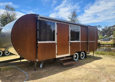 Rustic brown caravan with a modern design parked near a scenic waterway, available at Stawell Hotel Motel Accommodation. Ideal for a unique stay surrounded by nature