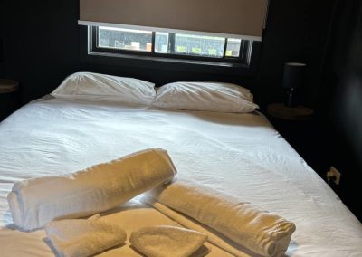 Cozy bedroom in Cabin 30 featuring a neatly made bed adorned with crisp white linens and fluffy pillows. Freshly folded white towels are artistically arranged on the bed, adding a touch of hospitality. The room is softly illuminated by a bedside lamp and has a window covered with a blind, allowing for natural light to filter in at Stawell Hotel Motel Accommodation