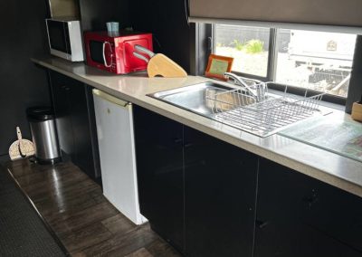 A modern kitchen area in Cabin 30 at Stawell Hotel Motel Accommodation featuring a sleek black design with a stainless steel sink, a red microwave, and a refrigerator. The countertop is equipped with essential kitchenware, and the large window allows natural light to illuminate the space, showcasing a tidy and functional environment for guests.