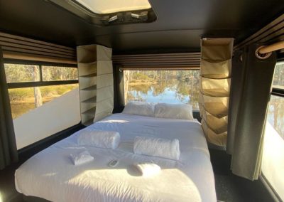Interior of a converted bus accommodation featuring a comfortable bed with large windows overlooking nature at Stawell Hotel Motel Accommodation