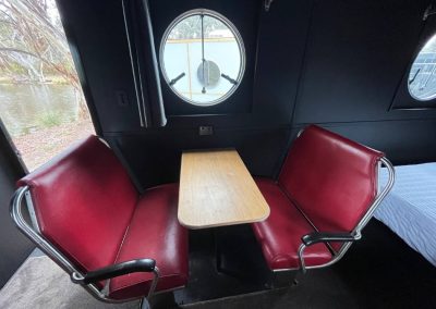 Red vintage-style booth seating with a wooden table in a caravan, next to a round window overlooking a lake at Stawell Hotel Motel Accommodation