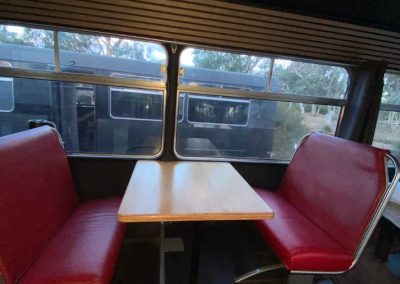 Retro-style dining area with red cushioned seats and a wooden table inside a converted caravan at Stawell Hotel Motel Accommodation, featuring large windows for scenic outdoor views