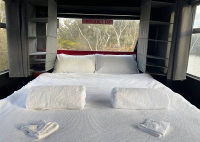 Interior view of a converted bus accommodation featuring a neatly made double bed with rolled towels, shelf storage, and large windows overlooking natural scenery at Stawell Hotel Motel Accommodation