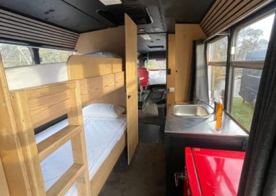 Interior of a converted bus with bunk beds, compact kitchenette featuring a red fridge and sink, leading to a dining area with red retro-style seats at Stawell Hotel Motel Accommodation