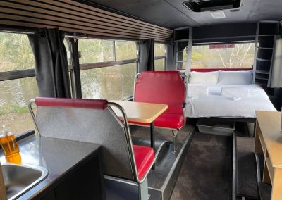 Interior view of a converted bus featuring a cozy bed at the rear, retro red dining booth with a table, and a compact kitchenette with a sink, overlooking a scenic riverside at Stawell Hotel Motel Accommodation