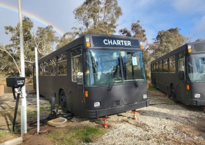 Exterior view of a converted charter bus at Stawell Hotel Motel Accommodation, parked outdoors with a rainbow in the background and another similar bus nearby, set against a backdrop of trees