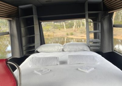 Interior of a converted charter bus at Stawell Hotel Motel Accommodation, featuring a neatly made double bed with views of the surrounding trees and water through large windows