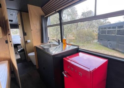 Kitchen area inside a converted charter bus at Stawell Hotel Motel Accommodation, featuring a sink, a bright red mini-fridge, and a view through the large windows of another bus and greenery outside