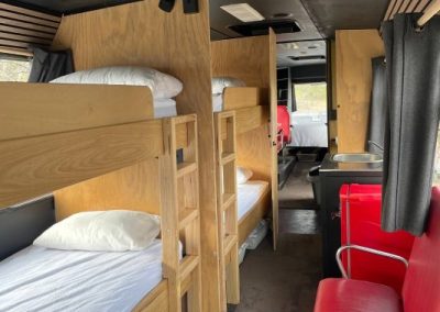 Interior of a converted charter bus at Stawell Hotel Motel Accommodation featuring custom wooden bunk beds, a red chair, and a table, with a view toward the kitchen and living areas in the background