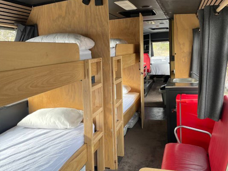 Interior of a converted charter bus at Stawell Hotel Motel Accommodation featuring custom wooden bunk beds, a red chair, and a table, with a view toward the kitchen and living areas in the background